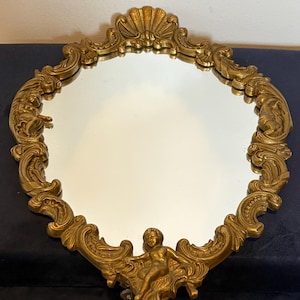 May include: An oval wall mirror featuring an ornate gold-colored frame with intricate scrollwork, a shell motif at the top, and a sculpted figure of a cherub at the bottom. The reflective glass surface is surrounded by detailed relief carvings.