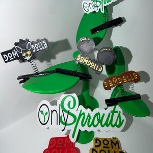 May include: A whimsical display featuring green, leaf-shaped decorations with attached black clips. Various labels with the text "Dom Dolla" and "Only Sprouts" are attached. Some labels are red, gold, and black.