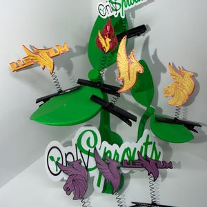 May include: A whimsical display featuring green leaf-shaped elements with attached black hair clips. Decorative accents include gold and purple winged figures, and the text "Only Sprouts" in a playful font. The design incorporates springs for added visual interest.