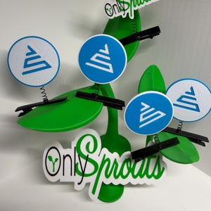 May include: A green, stylized plant with leaves and circular accents. The circles are white with a blue geometric design. The text "Only Sprouts" is visible in green and white. Black hair clips are attached to the leaves.
