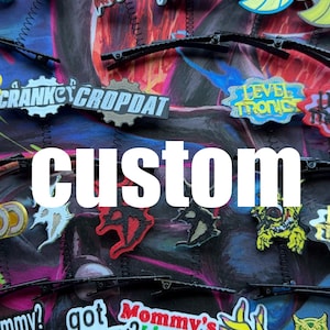 May include: An assortment of custom-made hair clips and patches with various designs and text. The word "custom" is prominently displayed in white. The clips are black, and the patches feature a variety of colors and graphics.