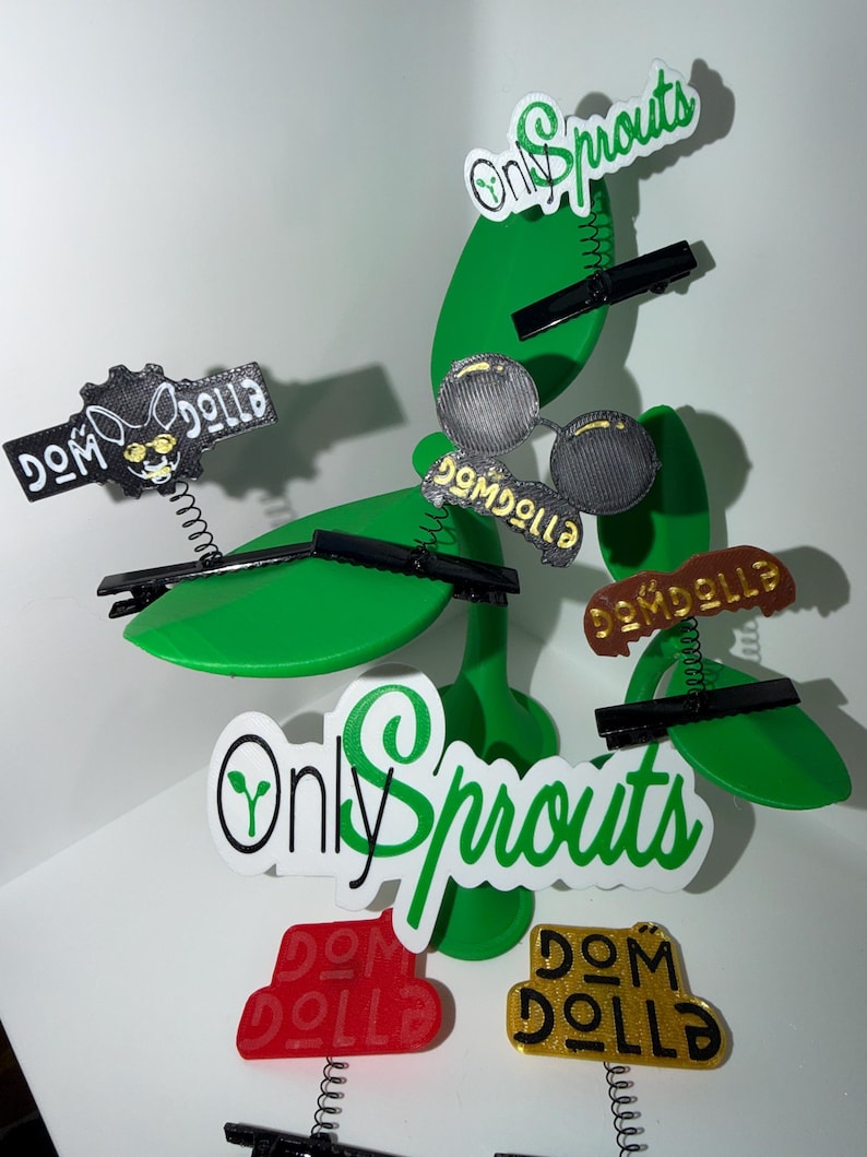 May include: A whimsical display featuring green, leaf-shaped decorations with attached black clips. Various labels with the text "Dom Dolla" and "Only Sprouts" are attached. Some labels are red, gold, and black.
