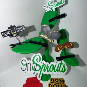 May include: A green, stylized plant sculpture with several decorative hair clips attached. The clips feature various designs, including the text "Only Sprouts" and "Dom Dolla". The clips are in black, red, gold, and silver.
