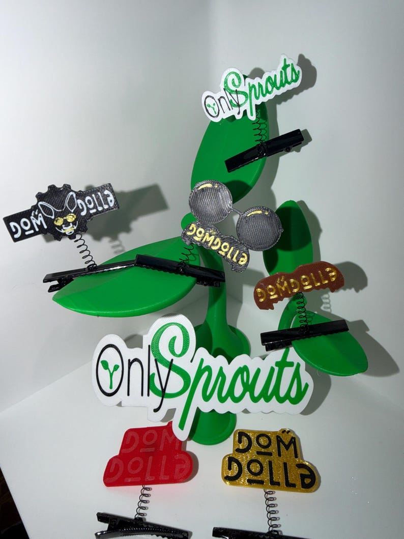 May include: A green, stylized plant sculpture with several decorative hair clips attached. The clips feature various designs, including the text "Only Sprouts" and "Dom Dolla". The clips are in black, red, gold, and silver.