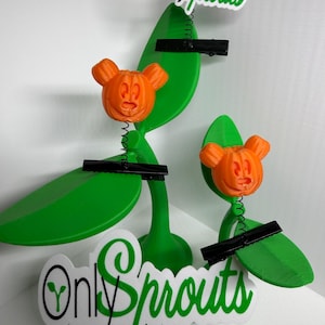 May include: Two green plant-shaped stands with orange pumpkin-shaped heads featuring mouse-like ears. Each stand has black metal clips attached to thin springs. The white and green text Only Sprouts is displayed on the base and top.