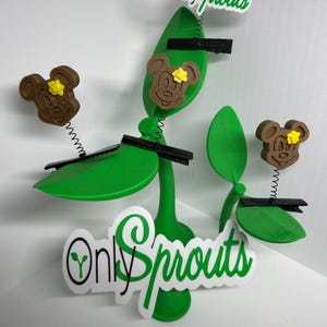 May include: Two green sprout-shaped stands featuring black hair clips attached to thin metal springs. Each spring is topped with a brown mouse-shaped head with a yellow flower. The white sign with green text reads Only Sprouts.