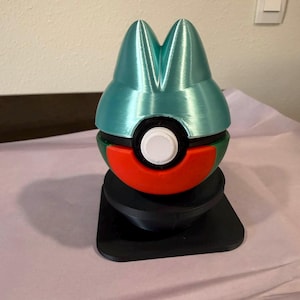 May include: A 3D-printed teal and red Poké Ball replica, featuring a black base and a white central button. The top section has a unique, pointed design. The object is displayed on a light pink surface.