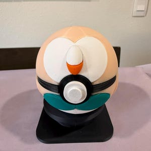 Könnte beinhalten: Ein kugelförmiges, 3D-gedrucktes Objekt, das einem Charakter mit pfirsichfarbenem Kopf, weißen Gesichtsmarkierungen, einem kleinen orangefarbenen Schnabel und einem türkisfarbenen Unterteil ähnelt. Es hat ein schwarzes Band mit weißem Knopf.