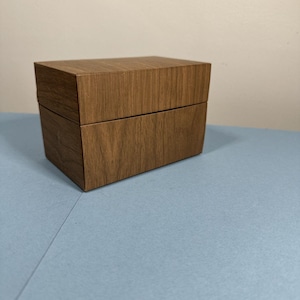May include: A rectangular wooden box with a hinged lid. The box has a wood grain pattern and a warm brown color. The box is sitting on a light blue surface.