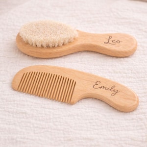 May include: A wooden baby brush and comb set. The brush has soft bristles and the comb has fine teeth. The brush is labeled "Leo" and the comb is labeled "Emily". Both are made of light-coloured wood.