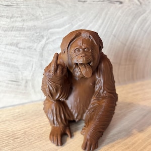 May include: A brown orangutan figurine with its tongue sticking out and middle finger raised. The sculpture has detailed fur texture and is posed in a seated position, conveying a humorous or defiant expression.