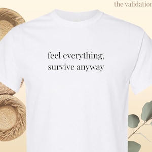 May include: A white short-sleeved t-shirt featuring the black text "feel everything, survive anyway" printed in a simple serif font across the center of the chest. The garment has a classic crew neckline and a standard fit with short sleeves.