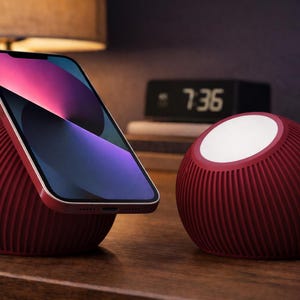 以下が含まれることがあります： リブ加工が施された赤い電話スタンドがスマートフォンを保持し、白い光のランプが隣にあります。デジタル時計は7:36を表示しています。これらのアイテムは木製の表面にあり、モダンな美学を作り出しています。