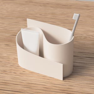 Puede incluir: Un soporte para cepillos de dientes beige con un diseño curvo, que contiene un tubo de pasta de dientes blanco y un cepillo de dientes gris. El soporte es de material liso y sólido, sobre una superficie de madera. Diseño moderno y minimalista.