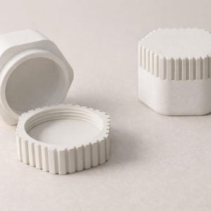 May include: Three white, hexagonal containers. One is open, revealing the interior. The lid is detached, showing a ridged edge. The other container is closed, with a ridged top. The containers are on a neutral background.
