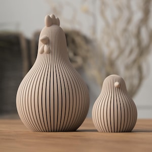 Könnte beinhalten: Zwei Holz-Hühnerfiguren, eine große und eine kleine, mit vertikalen Streifendetails. Das größere Huhn hat einen Kamm und einen Kehllappen. Die Figuren sind hellbraun und stehen auf einer Holzoberfläche.