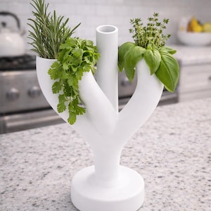 Könnte beinhalten: Ein weißer Kräutertopf aus Keramik mit drei Abschnitten, die jeweils frische Kräuter enthalten. Der Topf hat ein einzigartiges, baumartiges Design und steht auf einer gesprenkelten Arbeitsplatte. Rosmarin, Koriander und Basilikum sind sichtbar.