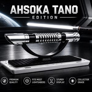 Pode incluir: Um sabre de luz prateado e preto com o texto "AHSOKA TANO EDITION" &eacute; exibido em um suporte preto. O sabre de luz est&aacute; em uma base branca com uma inser&ccedil;&atilde;o preta. A imagem tamb&eacute;m inclui um segundo sabre de luz no fundo.