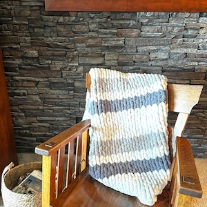 May include: A chunky knit blanket in shades of white, gray, and blue is draped over a wooden armchair. The blanket features horizontal stripes and a soft, textured appearance. The chair is made of dark wood.