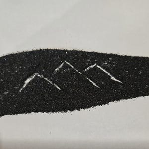 May include: A curved, elongated shape formed from fine black sand or granular material. Within the dark texture, two distinct mountain peak outlines are created by negative space, revealing the light-colored surface underneath the sand.