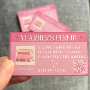 May include: A pink Yearner's Permit card with the text "YEARNER'S PERMIT" and "ALLOWS OWNER TO FALL IN LOVE WITH FICTIONAL CHARACTERS AND WORLDS." The card also features a stack of books and a QR code.