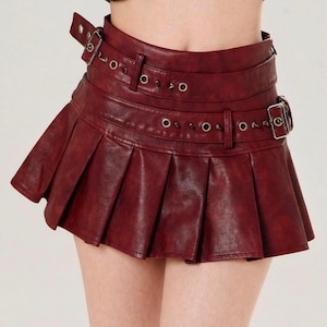 May include: Burgundy faux leather pleated mini skirt with multiple belt straps and silver-tone hardware. The skirt features a pleated design and a buckle closure. The skirt is a stylish addition to any wardrobe.