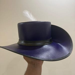 May include: A dark blue hat with a wide brim and a cylindrical crown. The hat features a black band around the crown and a single white feather. The hat is held up by a hand.