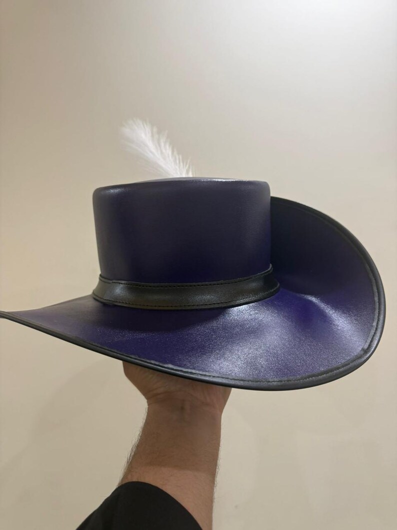May include: A dark blue hat with a wide brim and a cylindrical crown. The hat features a black band around the crown and a single white feather. The hat is held up by a hand.