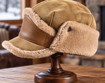 May include: A tan suede trapper hat featuring a smooth leather front panel on the brim and thick, cream-colored faux shearling lining along the edges and ear flaps. The hat has a rounded crown and a structured, short visor design.