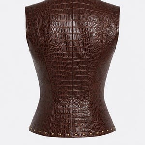 May include: A sleeveless vest in a dark brown color featuring a textured crocodile skin pattern throughout. The garment has a fitted silhouette with a straight neckline and a row of small, metallic gold-toned studs along the bottom hemline.