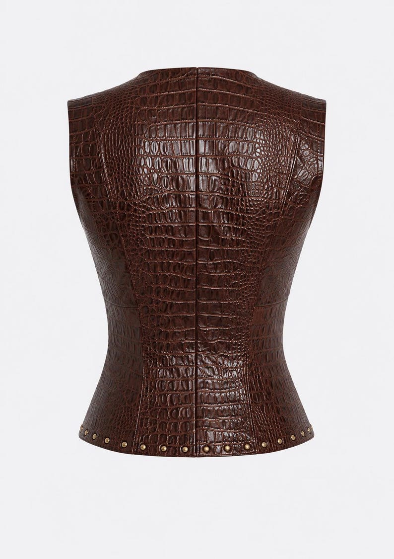 May include: A sleeveless vest in a dark brown color featuring a textured crocodile skin pattern throughout. The garment has a fitted silhouette with a straight neckline and a row of small, metallic gold-toned studs along the bottom hemline.