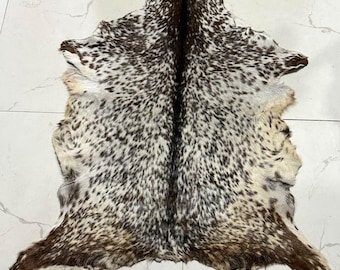 May include: A natural animal hide rug featuring a dense pattern of dark brown and black spots on a cream-colored background, with a prominent dark brown stripe running vertically down the center of the pelt's textured surface.