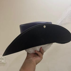 May include: A black tricorn hat with a wide brim, a dark blue crown, and a white feather. The hat is held up by a hand. A silver button is on the side of the hat.