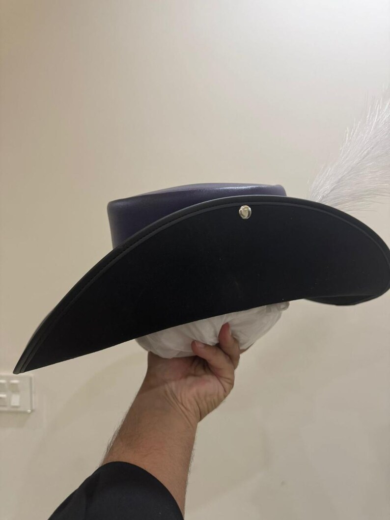 May include: A black tricorn hat with a wide brim, a dark blue crown, and a white feather. The hat is held up by a hand. A silver button is on the side of the hat.