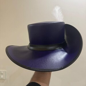 May include: A dark blue, wide-brimmed hat with a black band and a single white feather. The hat has a cylindrical crown and a broad, curved brim. The hat is held up against a neutral background.