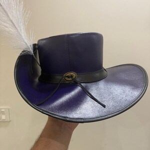 May include: A dark purple hat with a wide brim and a tall crown. The hat features a black band with a decorative buckle and a white feather. The hat is made of a shiny material and has a classic design.