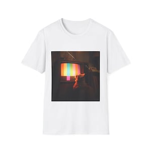 May include: White t-shirt featuring a retro image of a cat watching a vintage television. The TV screen displays a colorful test pattern. The cat is silhouetted against the screen's glow, creating a nostalgic and artistic design.