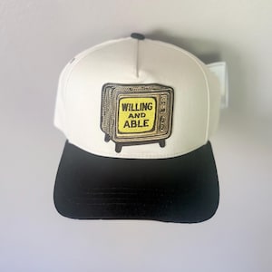 May include: A baseball cap with a cream-coloured crown and a solid black brim. Centered on the front is a rectangular patch featuring a vintage television set illustration with a yellow screen displaying the black text WILLING AND ABLE.