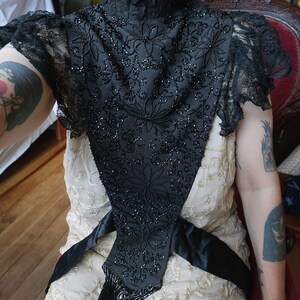 K&ouml;nnte beinhalten: Schwarzes, perlenbesetztes Vintage-Oberteil mit hohem Kragen und floralem Design. Das Oberteil hat aufwendige Perlenarbeiten und Spitz&auml;rmel. Ein schwarzer Satinsch&auml;rpe ist um die Taille gebunden, und das Oberteil hat Fransen.