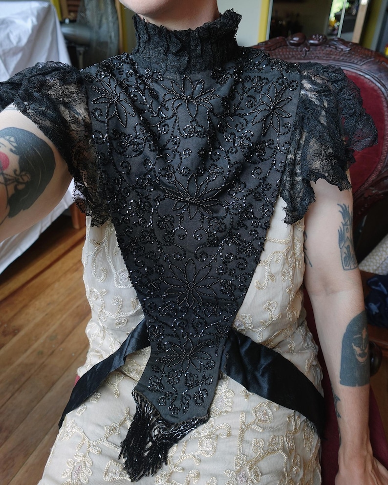 K&ouml;nnte beinhalten: Schwarzes, perlenbesetztes Vintage-Oberteil mit hohem Kragen und floralem Design. Das Oberteil hat aufwendige Perlenarbeiten und Spitz&auml;rmel. Ein schwarzer Satinsch&auml;rpe ist um die Taille gebunden, und das Oberteil hat Fransen.