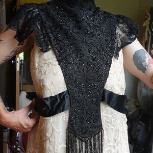 K&ouml;nnte beinhalten: Ein Vintage-Kragen und Schulterteil aus schwarzen Perlen mit floralen Designs und Fransen. Der Kragen wird &uuml;ber einem cremefarbenen Kleid mit gestickten Details und einem schwarzen Satinband getragen. Die &Auml;rmel sind aus Spitze.
