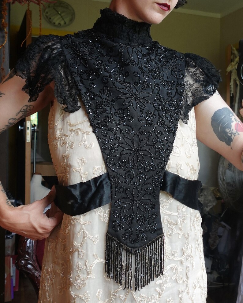 K&ouml;nnte beinhalten: Ein Vintage-Kragen und Schulterteil aus schwarzen Perlen mit floralen Designs und Fransen. Der Kragen wird &uuml;ber einem cremefarbenen Kleid mit gestickten Details und einem schwarzen Satinband getragen. Die &Auml;rmel sind aus Spitze.