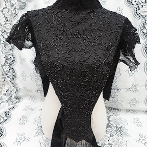 K&ouml;nnte beinhalten: Schwarze, mit Perlen bestickte Bolerojacke mit floralen Stickereien und Fransen. Die Vintage-Jacke hat einen hohen Kragen und kurze &Auml;rmel. Die aufwendige Perlenarbeit erzeugt ein detailliertes Blumenmuster auf dem Oberteil und dem R&uuml;cken.