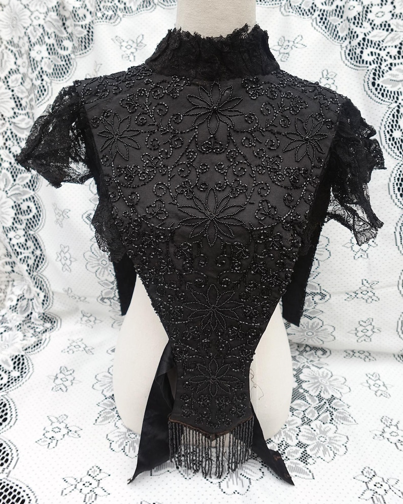 K&ouml;nnte beinhalten: Schwarze, mit Perlen bestickte Bolerojacke mit floralen Stickereien und Fransen. Die Vintage-Jacke hat einen hohen Kragen und kurze &Auml;rmel. Die aufwendige Perlenarbeit erzeugt ein detailliertes Blumenmuster auf dem Oberteil und dem R&uuml;cken.
