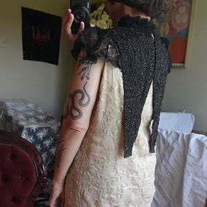 K&ouml;nnte beinhalten: Ein cremefarbenes Kleid mit einem komplizierten Blumenmuster wird mit einem schwarzen, bestickten Bolero kombiniert. Die Jacke hat einen hohen Kragen und kurze &Auml;rmel mit Spitzenbesatz. Das Kleid scheint aus einem weichen, flie&szlig;enden Stoff zu sein.