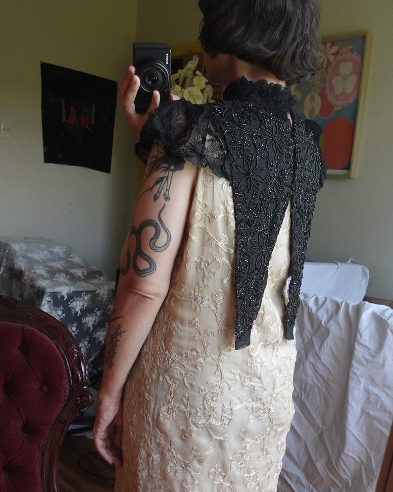 K&ouml;nnte beinhalten: Ein cremefarbenes Kleid mit einem komplizierten Blumenmuster wird mit einem schwarzen, bestickten Bolero kombiniert. Die Jacke hat einen hohen Kragen und kurze &Auml;rmel mit Spitzenbesatz. Das Kleid scheint aus einem weichen, flie&szlig;enden Stoff zu sein.