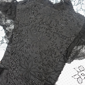 K&ouml;nnte beinhalten: Nahaufnahme eines schwarzen Textils mit aufwendiger floraler Perlenarbeit. Das Design zeigt gro&szlig;e, stilisierte Blumen und wirbelnde Muster, alles in schwarzen Perlen. Die R&auml;nder sind mit zarter schwarzer Spitze besetzt, was eine Vintage-&Auml;sthetik erzeugt.