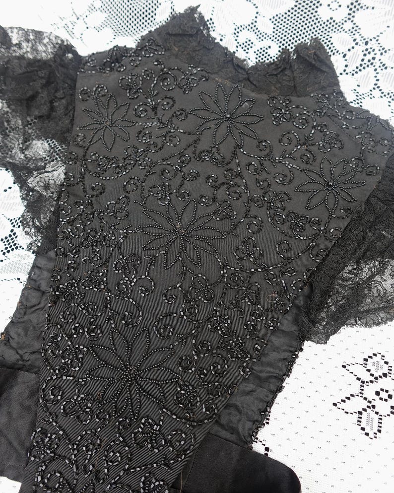 K&ouml;nnte beinhalten: Nahaufnahme eines schwarzen Textils mit aufwendiger floraler Perlenarbeit. Das Design zeigt gro&szlig;e, stilisierte Blumen und wirbelnde Muster, alles in schwarzen Perlen. Die R&auml;nder sind mit zarter schwarzer Spitze besetzt, was eine Vintage-&Auml;sthetik erzeugt.