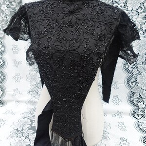 K&ouml;nnte beinhalten: Schwarzes Vintage-Oberteil mit Perlenbesatz, floraler Stickerei und Fransen. Das Oberteil hat einen hohen Kragen und kurze &Auml;rmel mit Spitzensaum. Die aufwendige Perlenarbeit erzeugt einen strukturierten, eleganten Look. Das Oberteil wird auf einer Schaufensterpuppe pr&auml;sentiert.