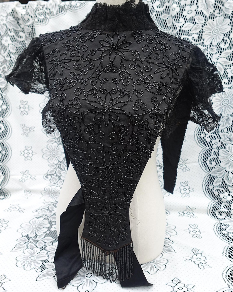 K&ouml;nnte beinhalten: Schwarzes Vintage-Oberteil mit Perlenbesatz, floraler Stickerei und Fransen. Das Oberteil hat einen hohen Kragen und kurze &Auml;rmel mit Spitzensaum. Die aufwendige Perlenarbeit erzeugt einen strukturierten, eleganten Look. Das Oberteil wird auf einer Schaufensterpuppe pr&auml;sentiert.